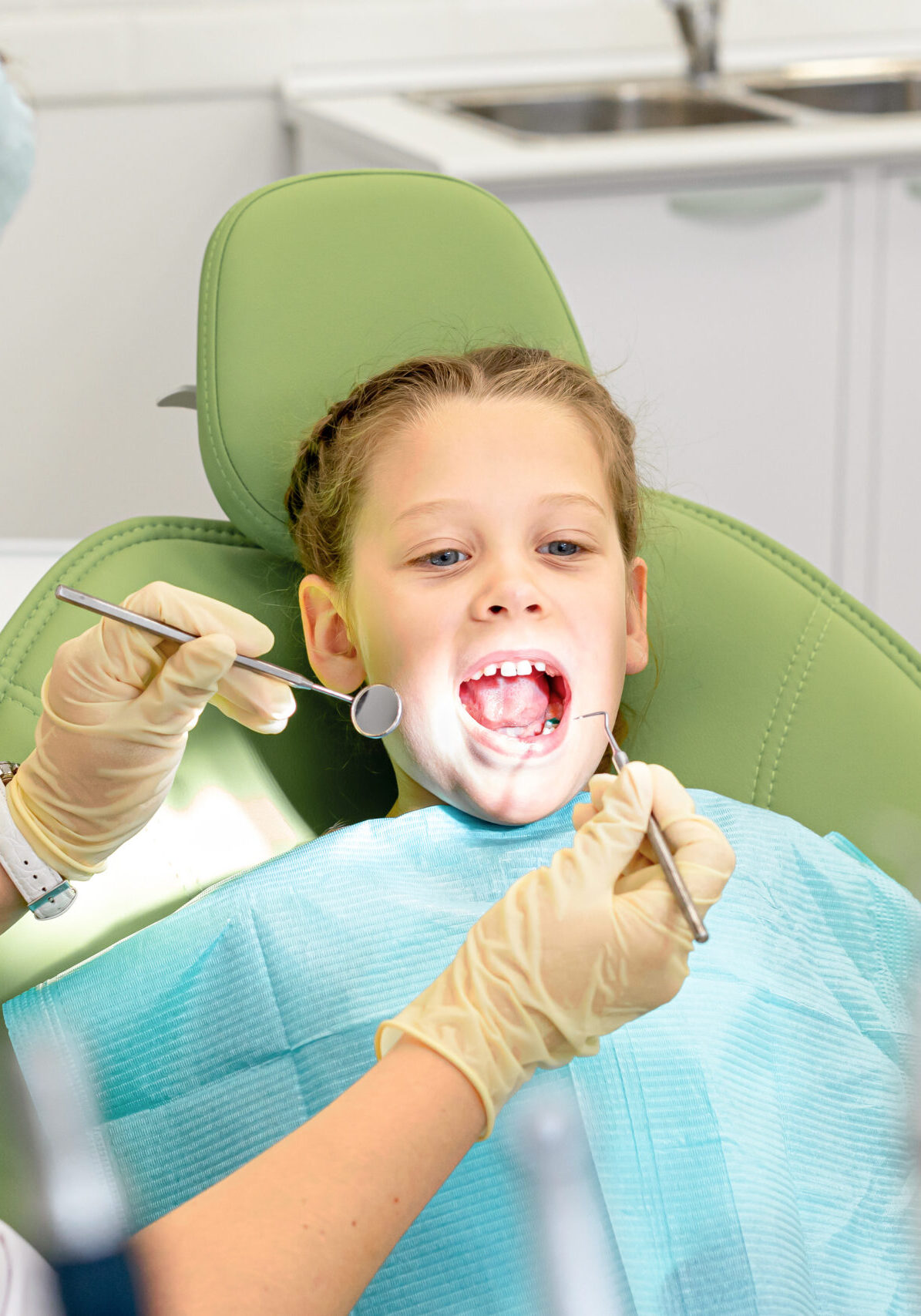 Little cute girl is sitting in dental chair and opening mouth in clinic, office. Doctor is preparing for examination of child teeth with instruments
