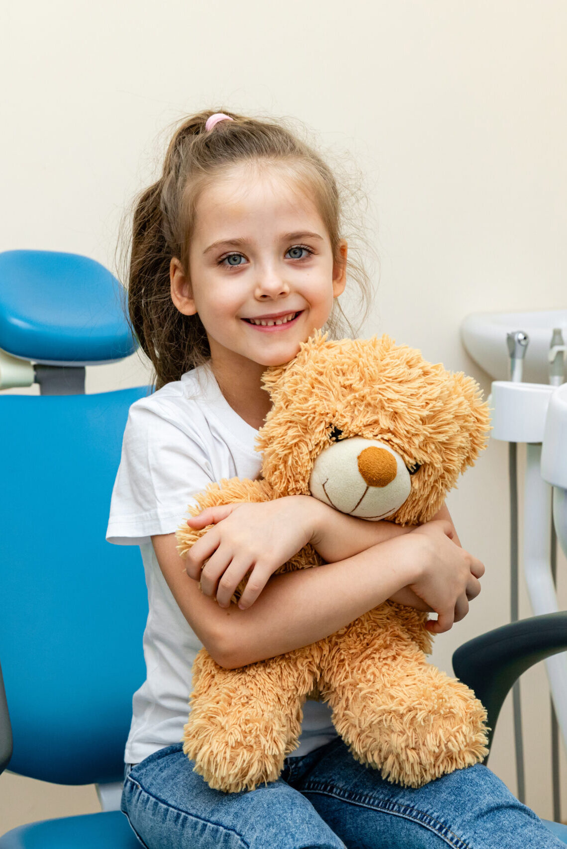 Happy little girl having dentist's appointment in modern clinic. Little patient, adorable girl sitting in dentist's chair during appointment in pediatric dentistry clinic and hugging her teddy bear.