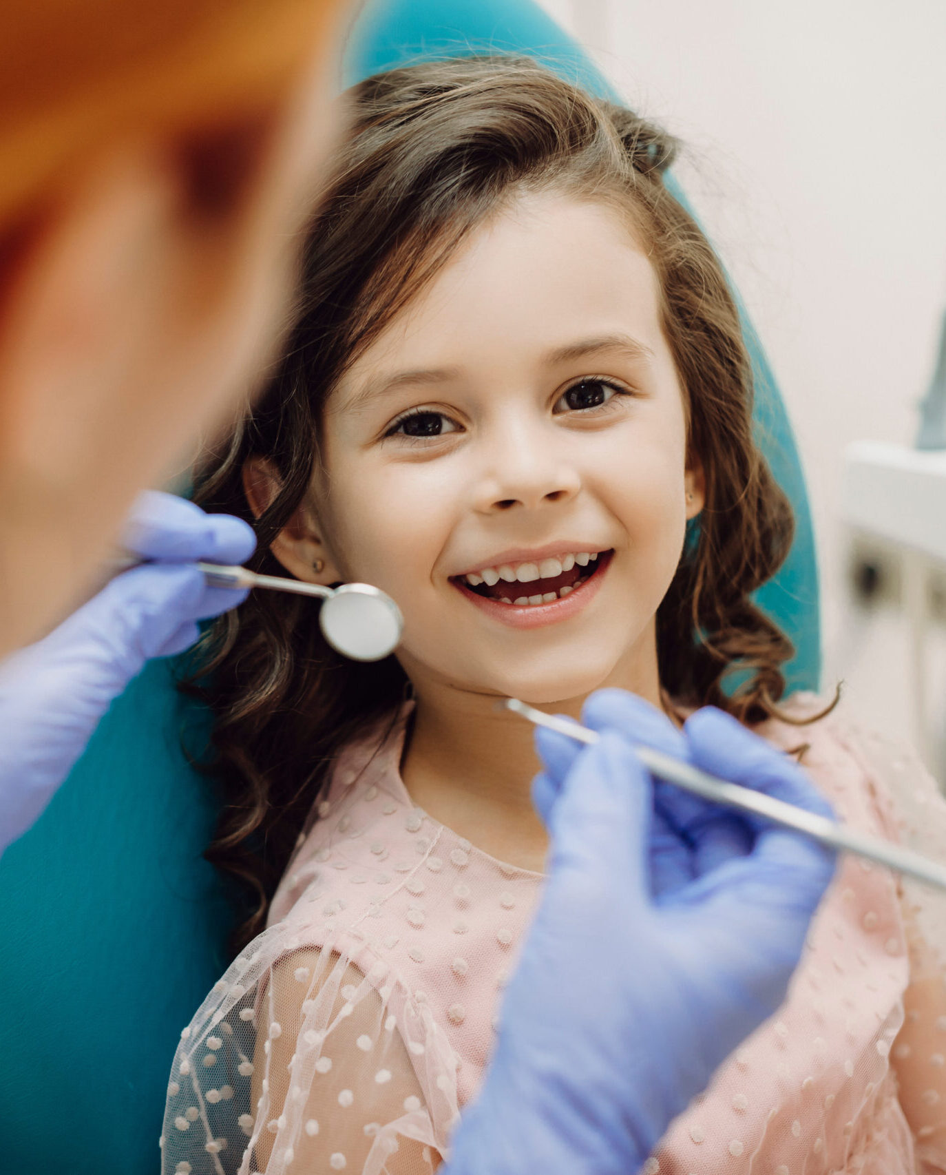 Portrait of a cute little girl laughing looking at camera sitting in stomatology seat while pediatric stomatologist is ready to do teeth examination.