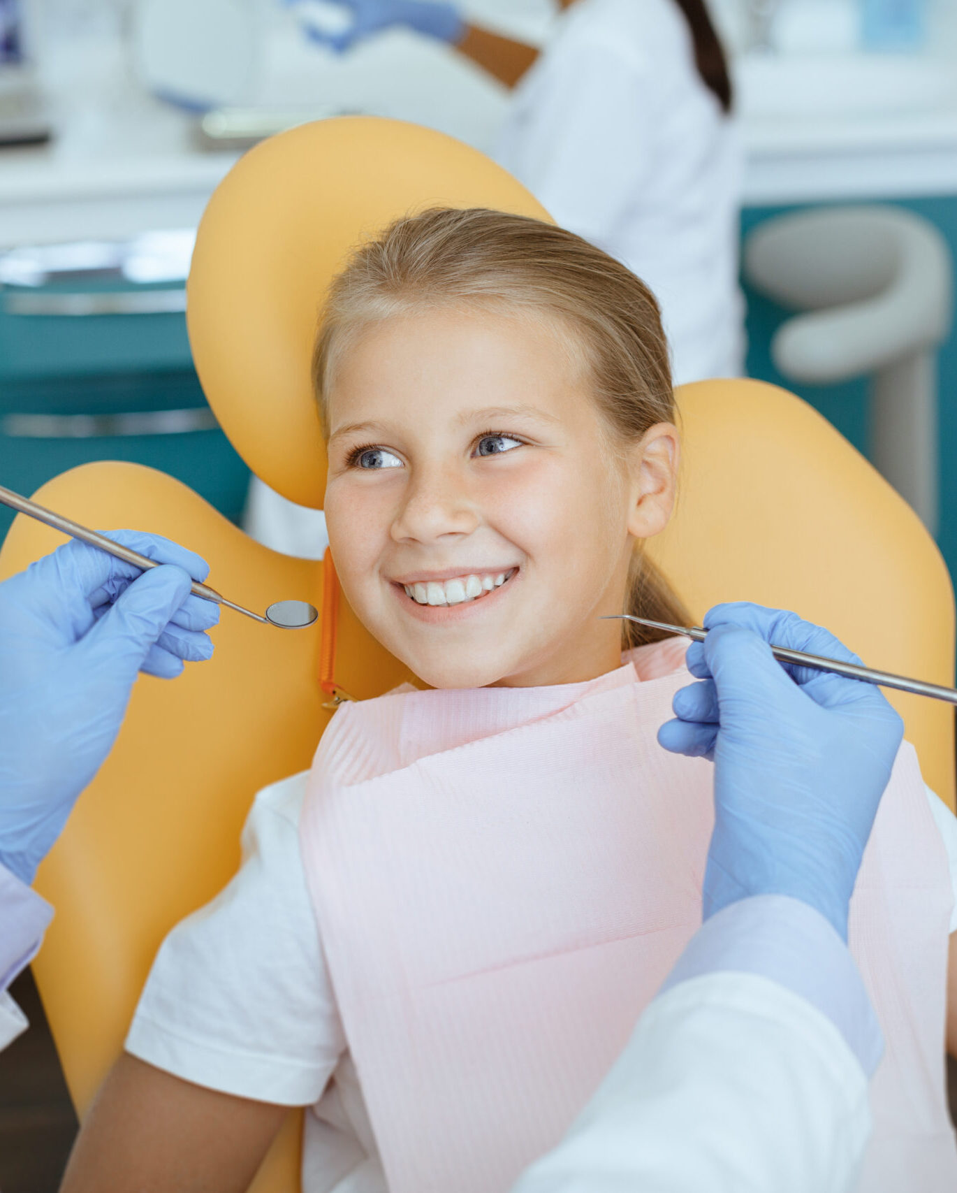 Fearless visit to doctor and dental treatment for child. Smiling girl in medical chair looks at dentist man in white coat, protective mask and rubber gloves with tools in his hands, and not afraid