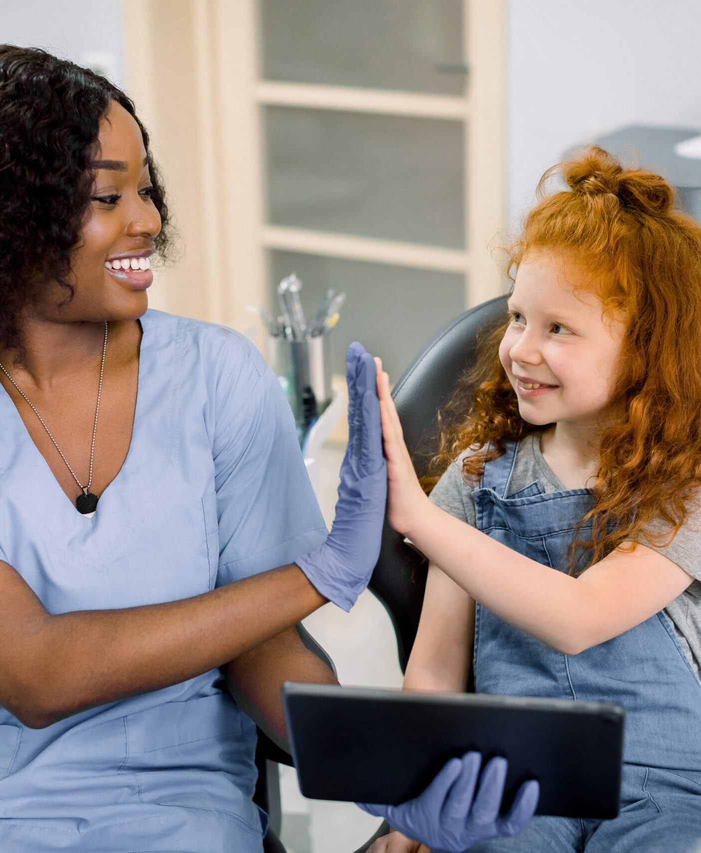 Medicine, pediatric dentistry and oral care concept. Happy female African dentist and curly red haired kid girl, having fun and giving high five, while holding tablet pc computer.