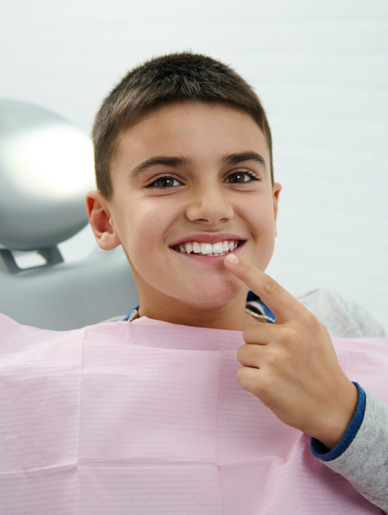 Happy boy holds finger near his mouth, looks at camera, smiles with beautiful toothy smile after receiving dental treatment in dentistry clinic. Oral hygiene, early prevention teeth diseases concept