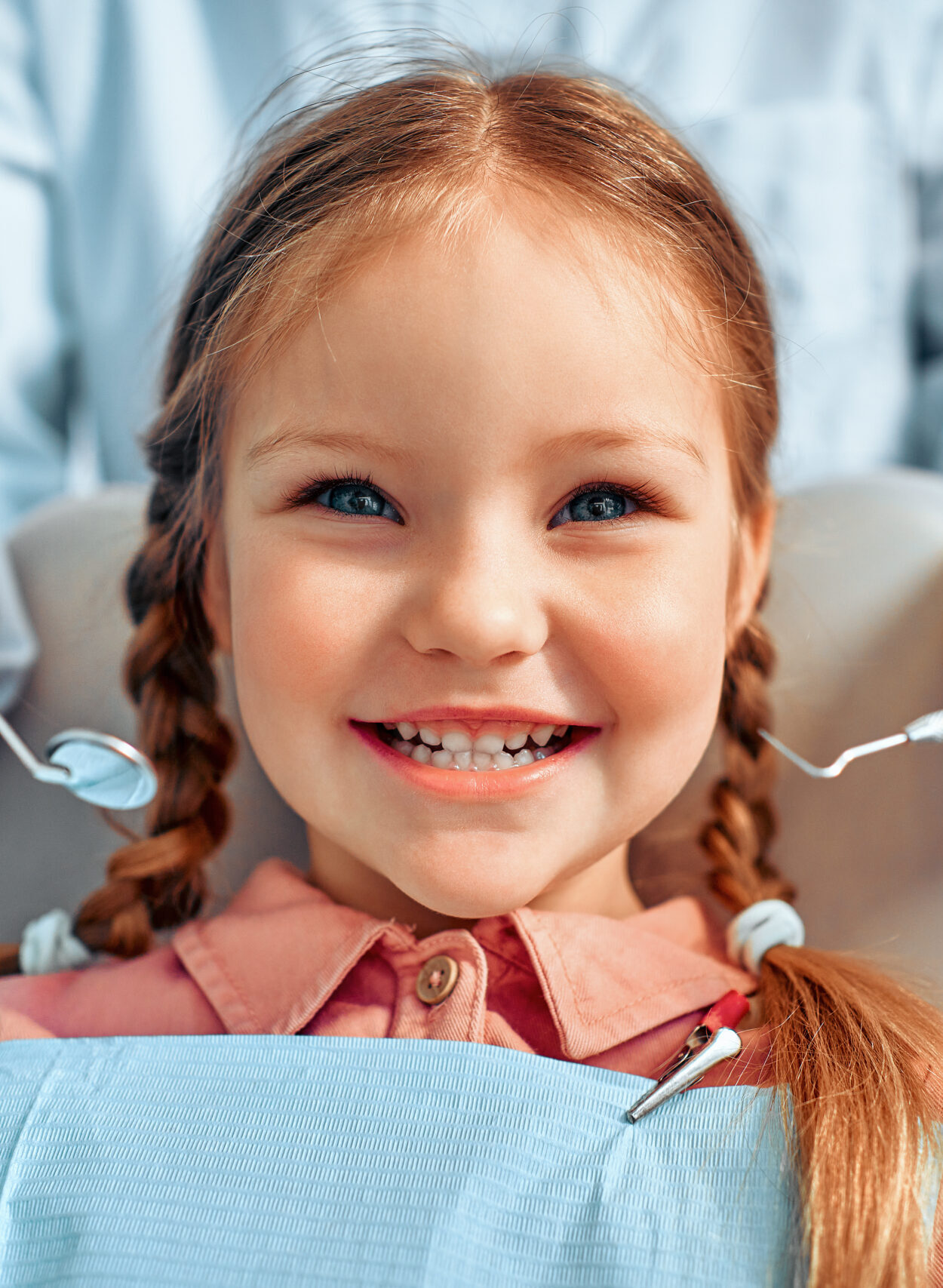 Cropped portrait of little girl with pigtails hair sitting in dental chair looking at camera and smiling. Behind, a doctor in gloves holds examination tools.Children's dentistry.