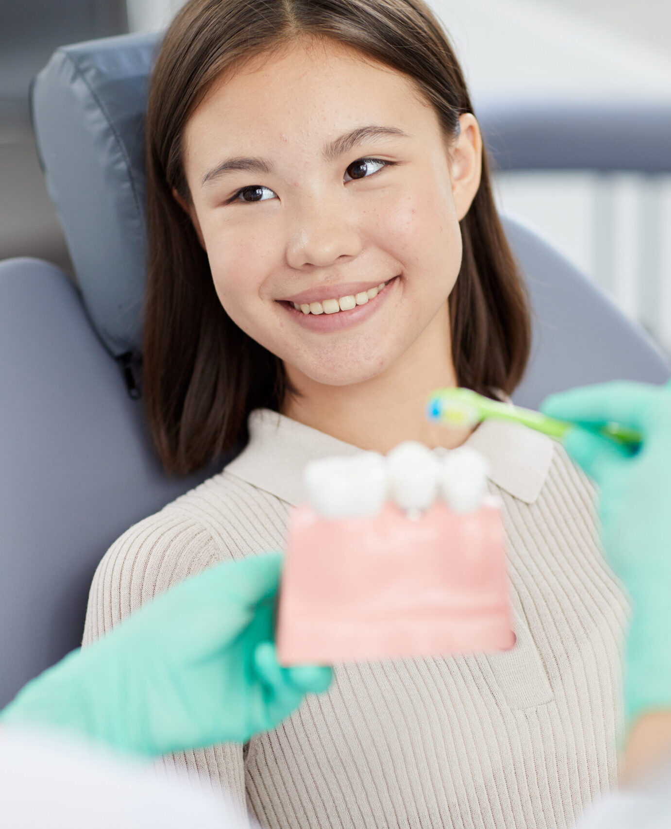 Portrait of smiling Asian girl sitting in dental chair and listening to dentist holding tooth model, copy space