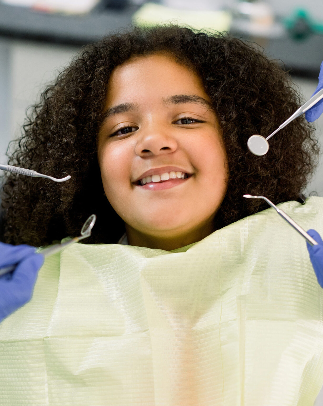Charming little African American girl afro hair, sitting in dental chair, smiling and looking at camera during medical treatment at modern dental clinic. Hands of two dentists with dental tools