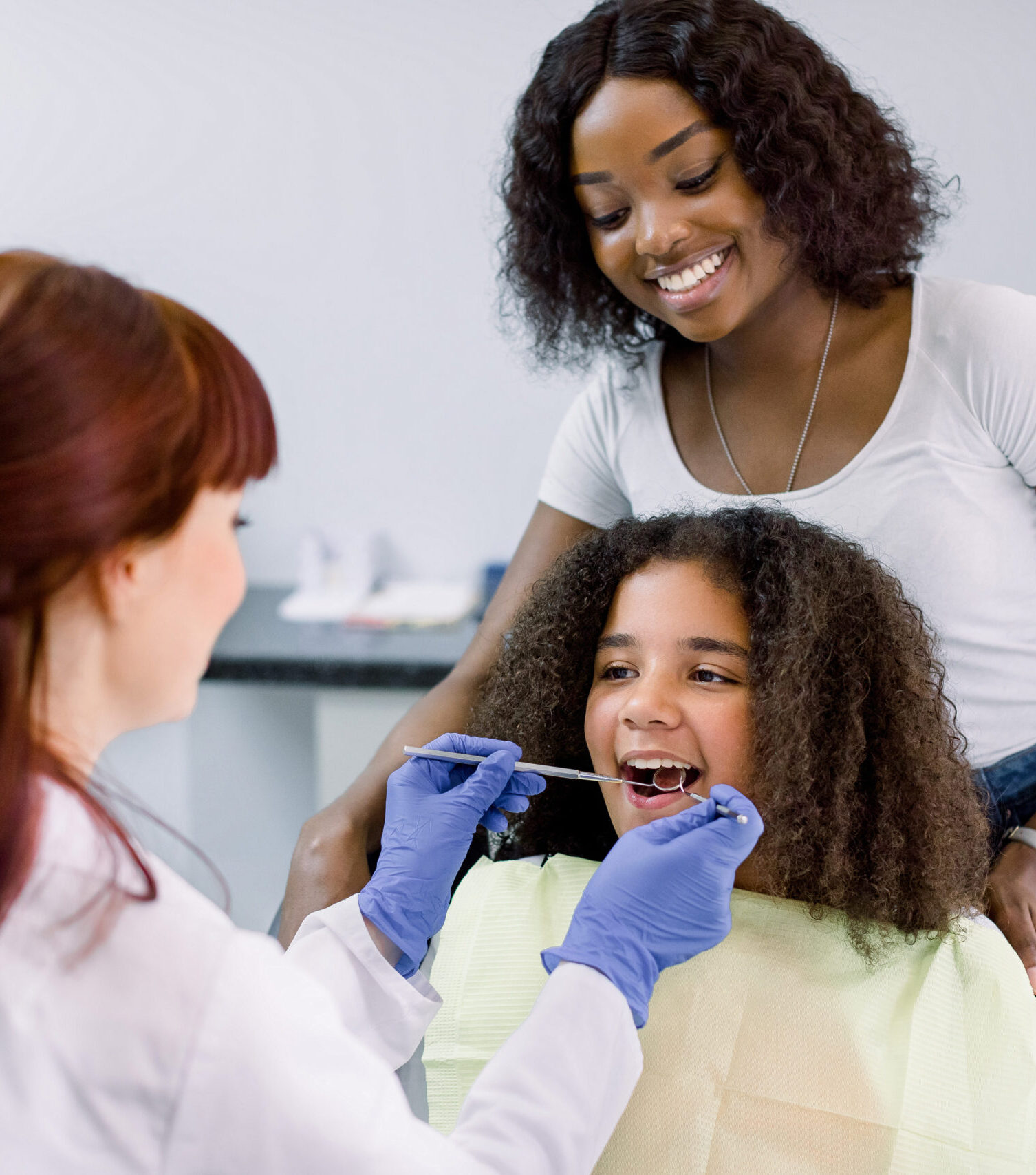 Dental treatment without fear. Female dentist in whilte uniform and gloves, examining teeth of little African American girl, while her mother supporting her behind, at modern pediatric dental clinic.