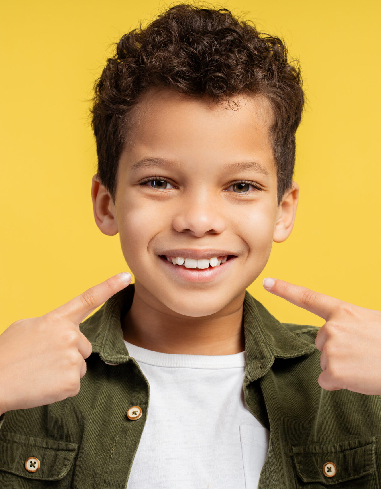 Handsome, smiling African American little boy pointing fingers at teeth standing isolated on yellow background. Dental care concept