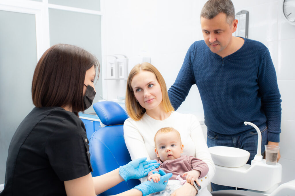 Young parents and their daughter visit a dentist to examine milk teeth. Baby at the first appointment with the dentist. Inspection of the formation of jaws, eruption of milk teeth. Family trip to the dentist