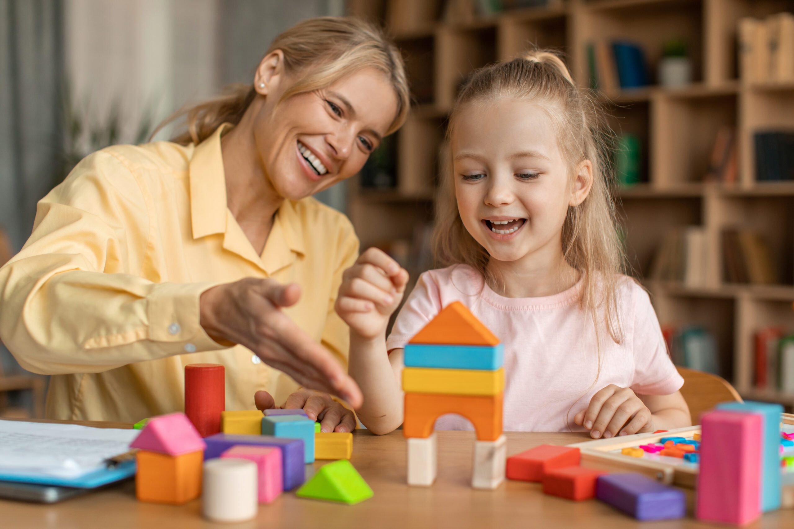 Cute little girl and cheerful child development specialist playing with colorful wooden bricks, sitting at table and making pyramid together, smiling